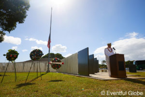 Photo of Pearl Harbor Day Memorial Ceremony -- military official giving a speech in front of a memorial and flag at half mast